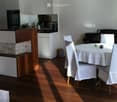 A modern dining area with a white tablecloth-covered table, six chairs, a piano, and a small kitchen area with cabinets and a faucet.
