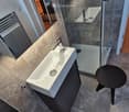 Modern bathroom with a white sink, chrome faucet, black stool, and a glass-enclosed shower area.