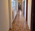 A long, narrow hallway with tiled flooring and white walls, featuring several doors and a visible bathroom at the end.