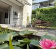 Garden area with a pond featuring pink lotus flowers, a patio with a table and chairs, and surrounding greenery.