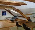 Open-plan living and dining area with exposed wooden beams, hardwood flooring, a black leather sofa, a dining table with chairs, a bookshelf, and a small kitchen area with cabinets.