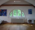 Bright attic room with wooden floors, large arched window, white radiator, bookshelf, small table, and paintings on the walls.