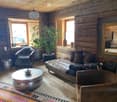 Cozy living room with wooden walls and floors, a large sofa, coffee table, armchair, floor lamp, and a potted plant near the window.