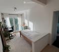 Bright bedroom with a wooden bed, desk, chair, laptop, and a plant near the window. The room features white walls, wooden flooring, and large windows with blinds.