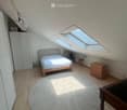 Cozy attic bedroom with wooden flooring, a round shaggy rug, a single bed, a wardrobe, a small table, and potted plants near the skylight windows.