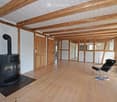 Open-plan space with exposed wooden beams, wooden flooring, and a black wood-burning stove. The room is unfurnished, with only a single black chair and a few books visible.