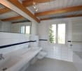 Bright bathroom with white tiled walls, two sinks, a toilet, a bathtub, and a large mirror. The room features wooden beams on the ceiling and a window with blinds.