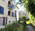 Multi-story residential building with blue shutters, balconies, and a small garden area with flowers and a lamp post.