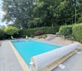 A rectangular swimming pool with a retractable cover, surrounded by a paved patio and lush greenery, including trees and shrubs. Two lounge chairs are placed near the pool, and a small staircase leads up to a higher section of the garden.