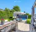 A well-maintained outdoor patio area with a wooden dining table, wicker chairs, a large umbrella, and a swimming pool in the background, surrounded by greenery and neatly trimmed hedges.