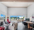 Modern living room with large windows offering scenic views, featuring a fireplace, sofa, chairs, a dining table, and a television.