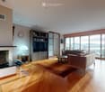 Modern living room with hardwood floors, large windows offering a scenic view, a fireplace, a sofa, a coffee table, a lamp, a bookshelf, and a small table with a stool.