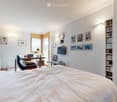 Bright bedroom with a large bed, desk with a laptop, comfortable chair, bookshelf, and wall-mounted TV. The room features large windows and modern decor.