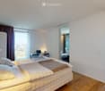 Modern bedroom with a large bed, wooden flooring, a desk with a laptop, a small table with a plant, and a mirror on the wall. The room has large windows with curtains and a view of the city.