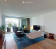 Modern living room with wooden flooring, large sliding glass doors leading to a balcony, a blue rug, a white armchair, a black leather chair, a small wooden side table, a television, and a lamp.
