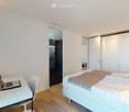 Modern bedroom with a white bed, laptop on a desk, white wardrobe, and a visible adjacent bathroom with dark tiles.