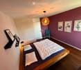 A modern bedroom with a wooden bed, white bedding, a blue armchair, a hanging geometric lamp, and several picture frames on the walls.