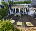 Wooden deck with a table, chairs, grill, and potted plants. Greenery and cushions are placed around the area.
