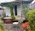 Garden area with a small shed, a table, chairs, a bench, and a cat resting on it. The shed has a door and is surrounded by greenery and flowers.