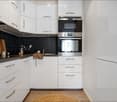 Modern kitchen with white cabinets, stainless steel oven, and black tiled backsplash. Includes a wooden cutting board, cup, and various kitchen utensils on the counter.
