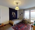 Cozy bedroom with a single bed, a patterned rug, a wooden chair, a small table, and a hanging lamp. A large mural of horses adorns one wall, and a sliding glass door leads to a balcony.