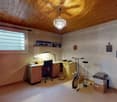 Cozy, well-lit room with wooden ceiling, large window, desk with chair, exercise bike, and storage boxes.