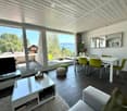 Modern open-plan living and dining area with large windows offering a scenic mountain view. Features a white coffee table, grey sofa, dining table with green chairs, and a mirror on the wall.
