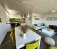 Open-plan living and dining area with modern furniture, including a white sofa, grey armchair, white coffee table, white dining table with green chairs, and a small kitchen area with white cabinets and a green backsplash.