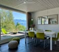 Modern dining room with white dining table, green chairs, two round stools, large mirror on wall, vase with flowers, and a view of mountains through large windows.