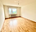 Empty room with wooden flooring, white walls, and a large window offering a view of the sea and surrounding landscape.