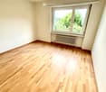 Empty room with wooden flooring, white walls, and a large window with a view of greenery. A radiator is visible beneath the window.