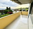 Balcony with a smooth white floor, yellow walls, and a striped awning. Offers a view of a suburban area with trees, buildings, and a body of water in the distance.