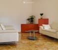 Two white leather sofas, glass coffee table, orange cabinet with plants, beige tiled floor, white walls, window with curtains