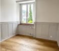 Empty room with wooden flooring, white walls, and a large window with a view of greenery outside.