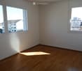 Empty room with wooden flooring and large windows offering a view of the city and surrounding buildings.