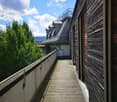Wooden balcony with metal railing, overlooking a green landscape and a building with a grey roof.