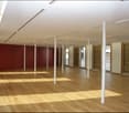 Empty room with wooden flooring, white pillars, and large windows with blinds. No furniture or equipment present.