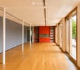 Empty room with wooden flooring, white walls, and large windows. Features include a red cabinet, white columns, and a sliding glass door leading to a balcony.
