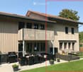 Modern two-story house with a balcony, patio seating area, and garden. Features include a dining set, outdoor furniture, and potted plants.
