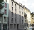 Multi-story residential building with a mix of modern and traditional architectural styles, featuring balconies, windows, and parking spaces on the street level.