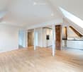 Bright, open attic space with wooden flooring, white walls, exposed brick pillar, and built-in kitchen cabinets. The room features large skylights and a staircase leading to another area.