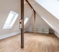 Empty attic room with wooden flooring, exposed wooden beams, and built-in white shelving unit. Skylight windows provide natural light.