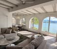 Bright living room with large arched windows overlooking a lake, featuring a grey sectional sofa, coffee table, armchair, dining area with table and chairs, and floor lamps.