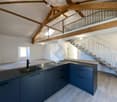 Modern kitchen with dark grey cabinets, a central island, and a staircase leading to an upper level. Exposed wooden beams on the ceiling.