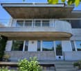 Modern two-story concrete house with large windows, a balcony, and a raised ground floor.