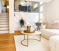 Modern living room with white walls, wooden flooring, a white sofa, a round coffee table with a gold frame, a white shag rug, a potted plant, and a vase with flowers on the coffee table. A staircase leads to an upper level with a railing.