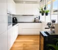 Modern kitchen with white cabinets, wooden flooring, and stainless steel appliances including an oven, coffee machine, and refrigerator. Potted plants are visible on the windowsill and counter.