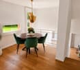 Dining area with a round wooden table, four green velvet chairs, a hanging gold pendant light, and a potted plant near the window.