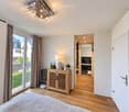Modern bedroom with wooden flooring, large window with curtains, bed, small wooden cabinet with decorative items, table lamp, and an open door leading to a modern bathroom with a sink, mirror, and cabinet.