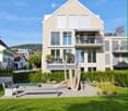 Modern two-story house with light beige exterior, large windows, and balconies. The front yard features a playground with a slide and wooden play structure, along with benches and greenery.